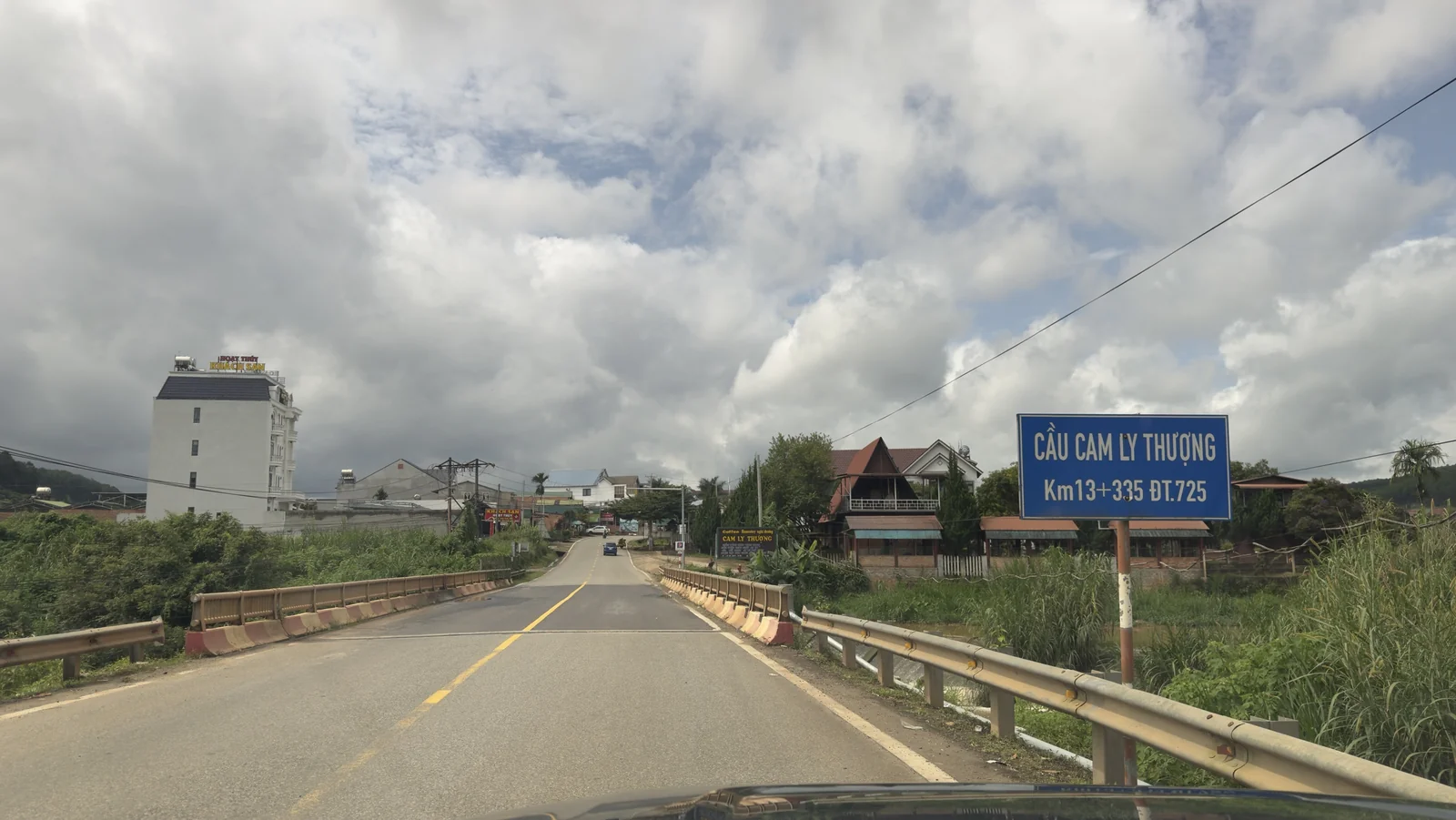 Cầu Cam Ly Thượng trên ĐT.725, biển tên màu xanh bên phải ghi Cầu Cam Ly Thượng Km13+335 ĐT.725, phía trước là xóm nhà và quán cà phê