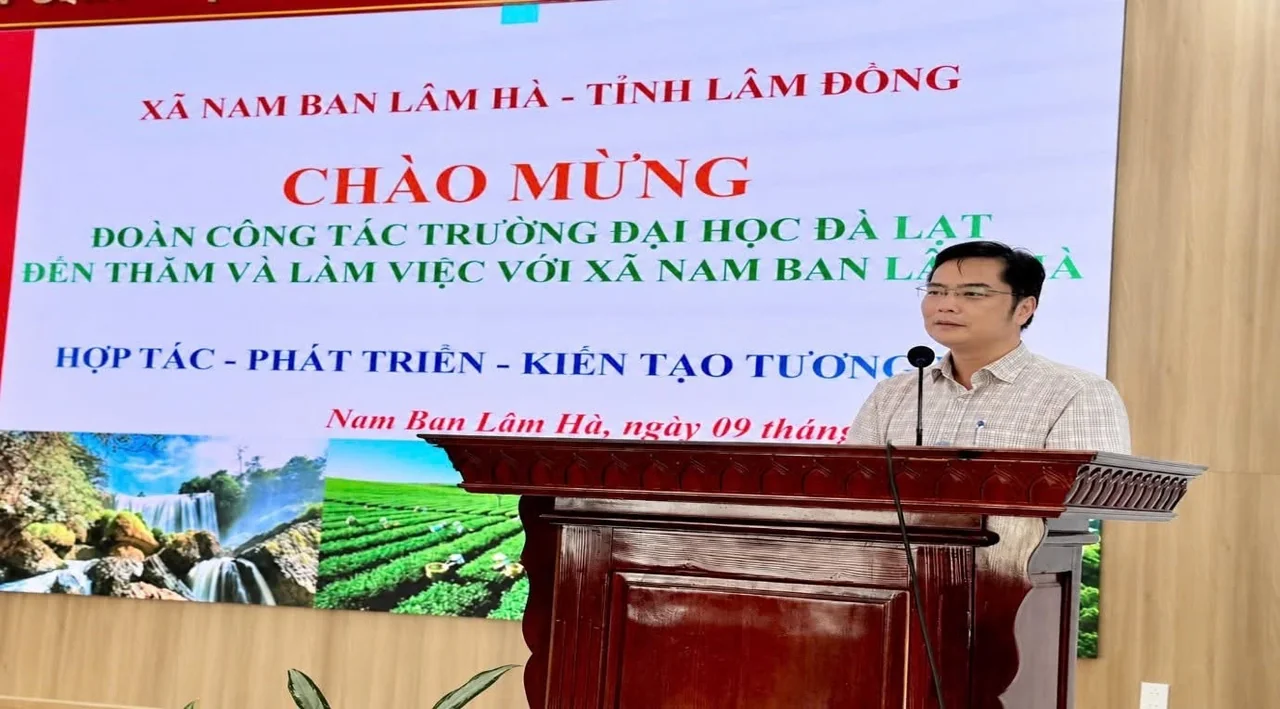 Buổi làm việc tại hội trường xã Nam Ban Lâm Hà, một người phát biểu tại bục, phía sau là màn hình chiếu dòng chữ chào mừng đoàn công tác Trường Đại học Đà Lạt