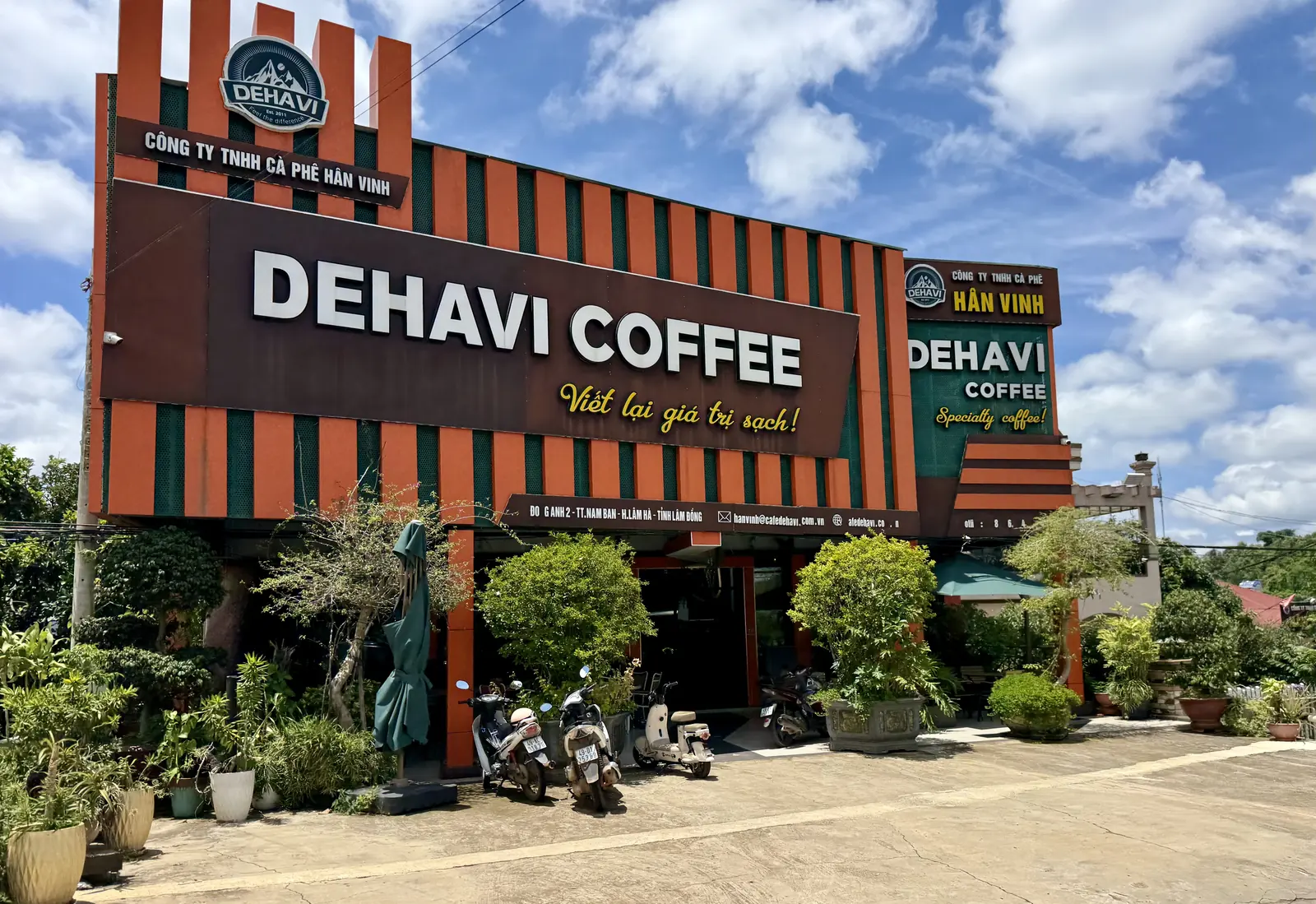 Mặt tiền cửa hàng Dehavi Coffee của Công ty TNHH Cà phê Hân Vinh ở Nam Ban, Lâm Hà — biển hiệu nâu và cam, cây cảnh cùng xe máy đỗ phía trước