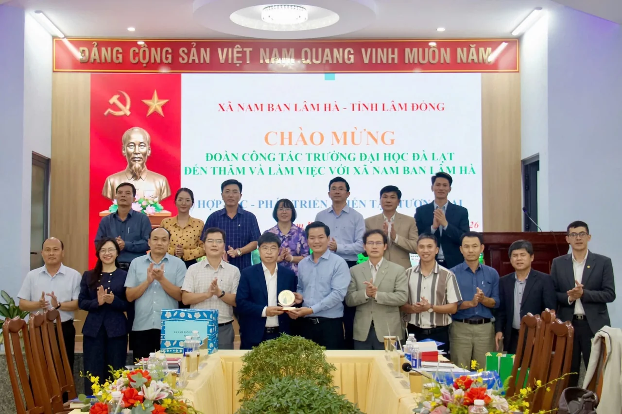 Hai đoàn đại biểu đứng chụp ảnh chung trước phông chào mừng trong hội trường xã Nam Ban Lâm Hà, ở giữa là nghi thức trao quà lưu niệm