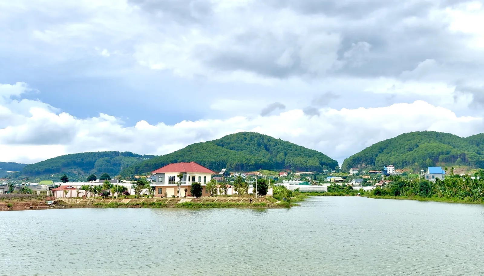 Hồ Từ Liêm ở Nam Ban nhìn từ bờ bên kia, có một căn nhà mái đỏ sát mép nước, phía sau là xóm nhà và đồi thông