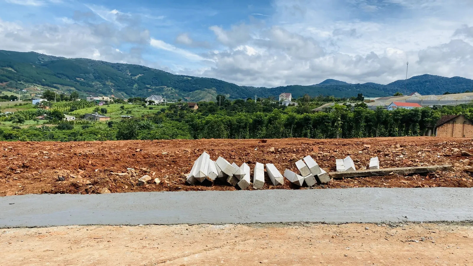 Đường bê tông vừa đổ chạy qua một lô đất đã san ở Nam Ban, bó vỉa bê tông xếp chờ lắp, phía sau là vườn cà phê và dãy đồi