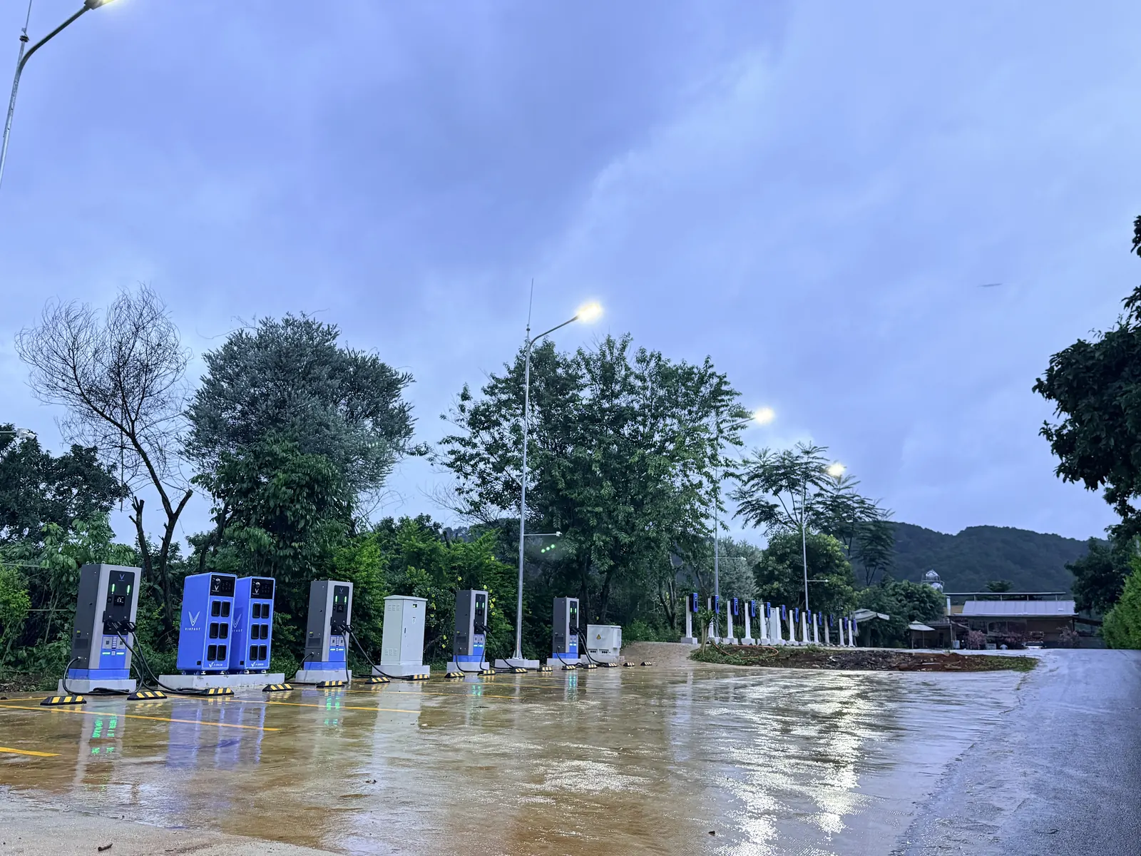 Trạm sạc VinFast nhiều trụ ở Nam Ban lúc chiều mưa, sân bê tông còn đọng nước, đồi phía sau