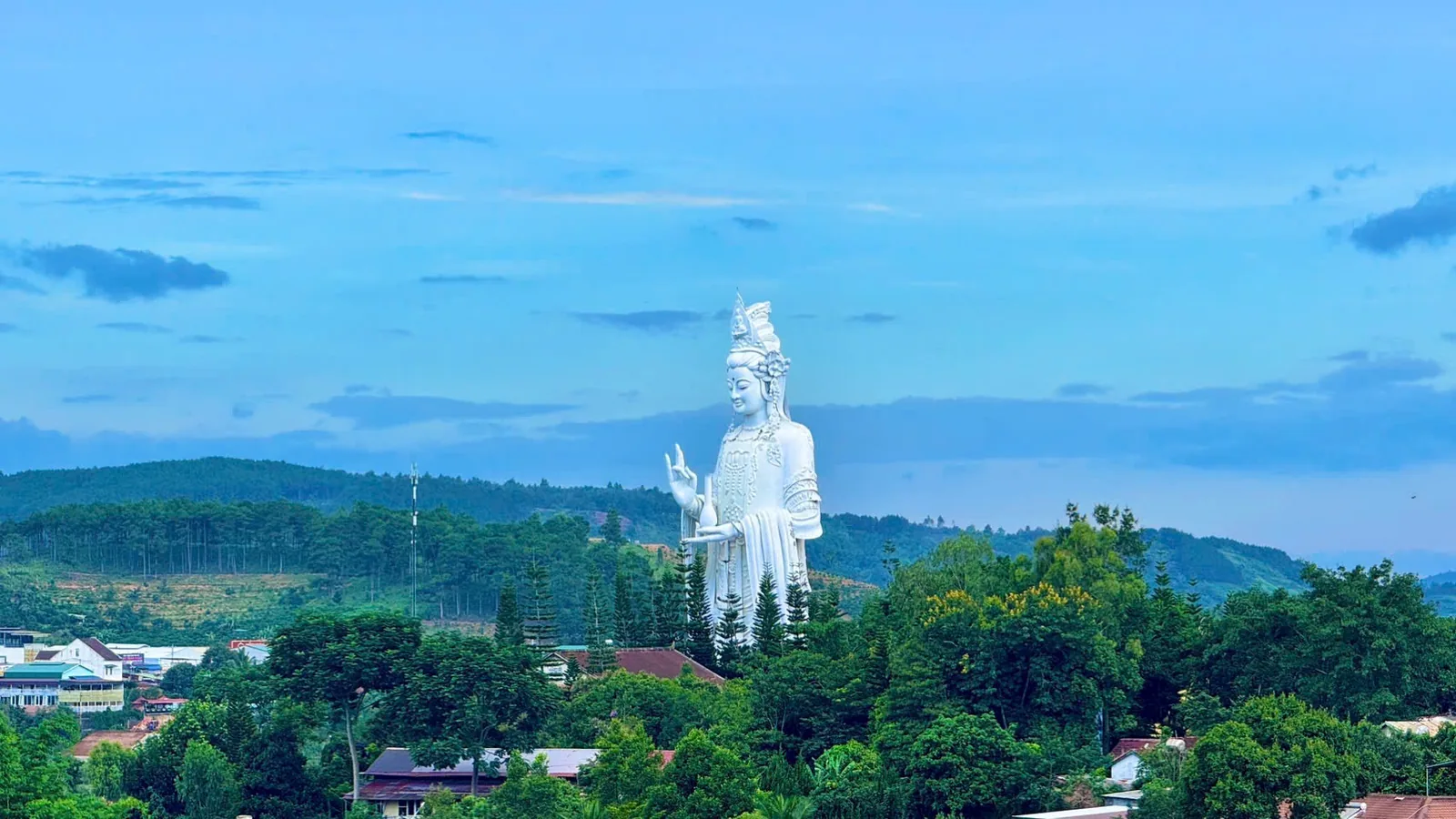 Tượng Quan Thế Âm chùa Linh Ẩn nhô cao trên mái nhà và hàng cây của khu dân cư Nam Ban, nền đồi xa và trời xanh